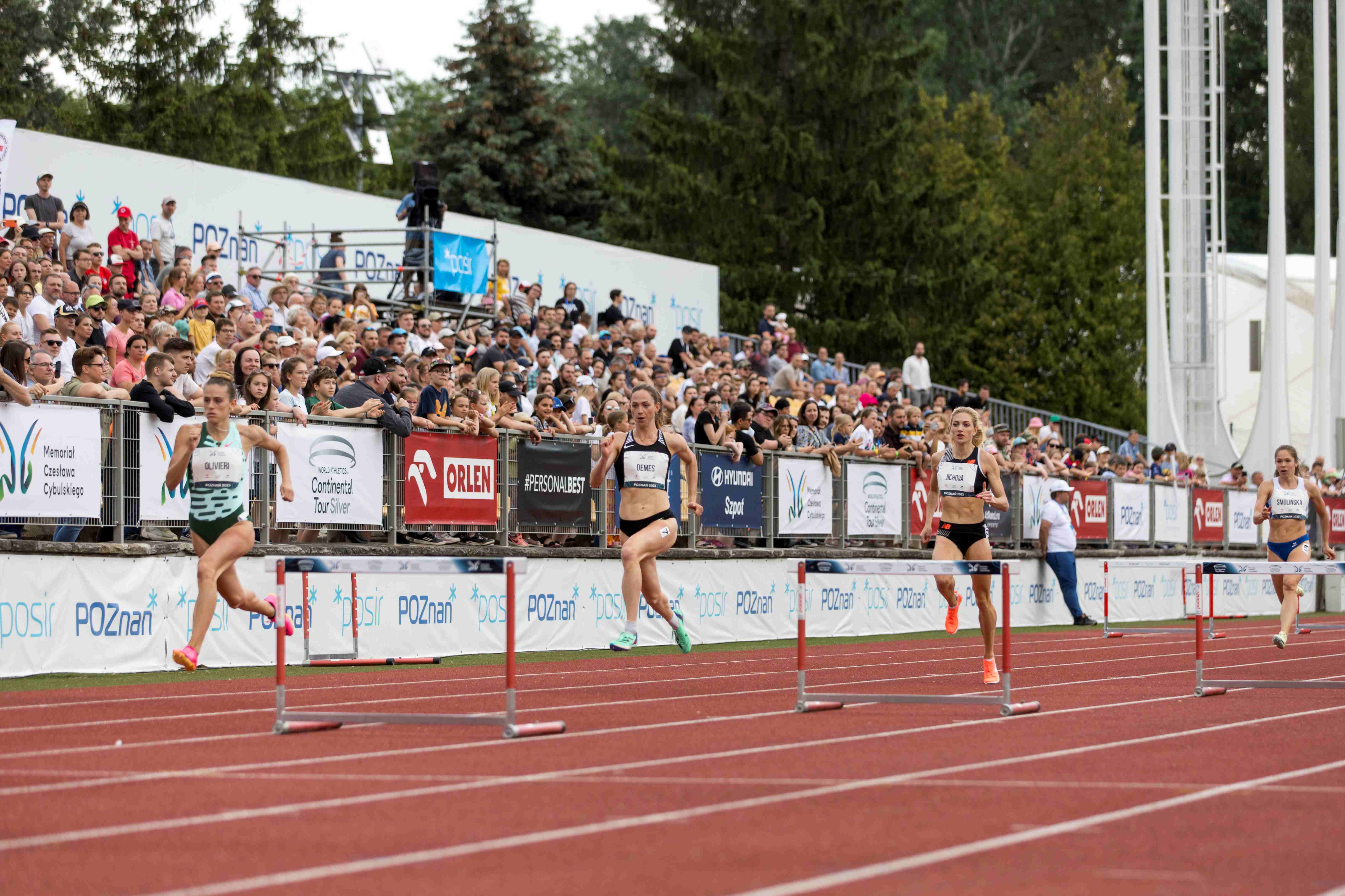 Hurdlers run in front of a grandstand full of spectators